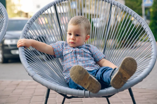 Niño jengibre vestido con jeans y camiseta azul sentado al aire libre en silla de mimbre . — Foto de Stock