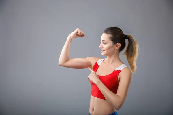 Hermosa mujer joven con estilo está apuntando a sus músculos y sonriendo, sobre fondo gris —  Fotos de Stock