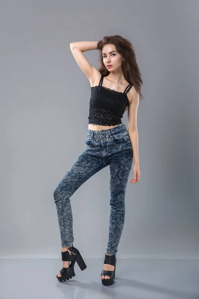 Ritratto di bella sexy modello donna con belle gambe lunghe in piedi indossando jeans denim nero lingerie top, posa guardando la fotocamera su sfondo grigio — Foto Stock