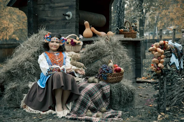 Adolescente assise dans du foin portant une couronne et un costume ukrainien national à côté du panier de fruits — Photo