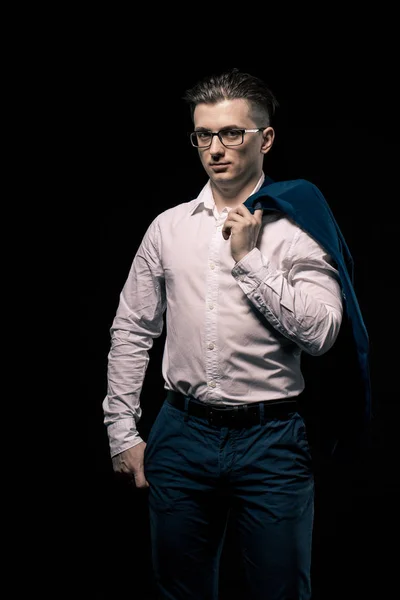 Homem de negócios elegante confiante com óculos olhando para a câmera sobre fundo estúdio preto. no ombro do homem segura uma jaqueta — Fotografia de Stock