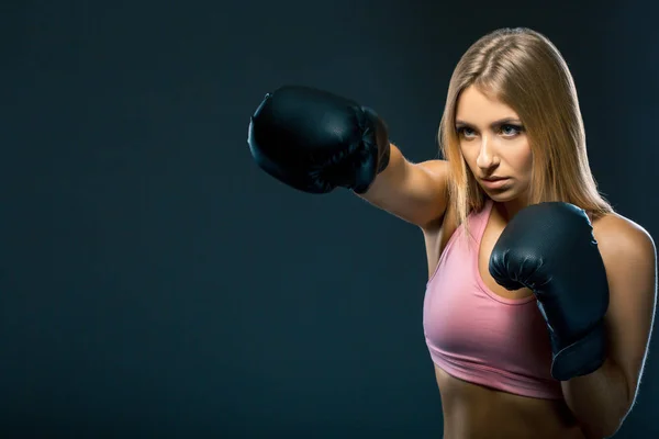 Woman in boxing gloves and sportswear strikes at workout on dark background. Copy space — Stock Photo, Image