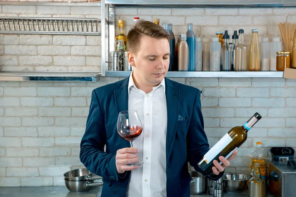 Sommelier apresentando uma garrafa de vinho tinto. Ele está de pé no interior da cozinha e segurando um copo . — Fotografia de Stock