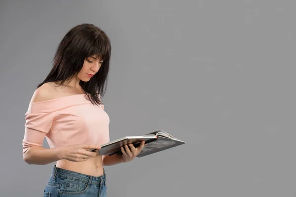Allvarlig attraktiv ung kvinna eller student som läser en bok som hon håller det i båda händerna stående mot en grå studio bakgrund — Stockfoto