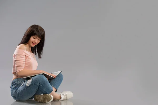 Молода жінка розслабляється на підлозі читає книгу на сірому фоні — стокове фото