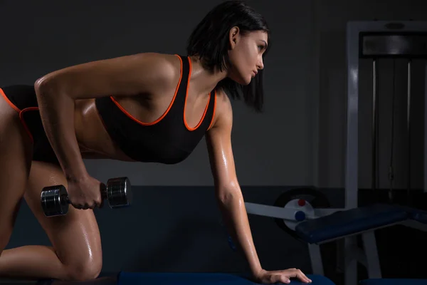 La fille dans la salle de gym s'accroupit avec un haltère, dans un bel uniforme de sport, sur un fond sombre . — Photo