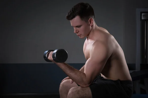 Knappe jonge man in korte broek, doen oefeningen voor de biceps, op een donkere achtergrond in de studio — Stockfoto