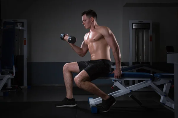 Knappe jonge man in korte broek, doen oefeningen voor de biceps, op een donkere achtergrond in de studio — Stockfoto