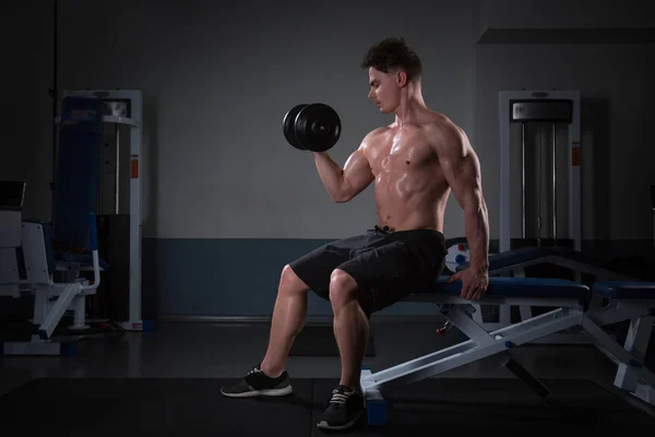 Knappe jonge man in korte broek, doen oefeningen voor de biceps, op een donkere achtergrond in de studio — Stockfoto