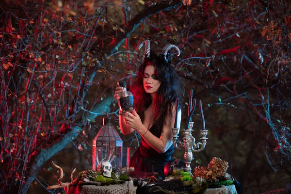 Bruxa de Halloween com chifres na cabeça segurando uma garrafa de poção no fundo da floresta — Fotografia de Stock