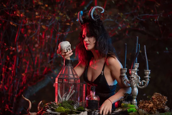 Belle sorcière avec des cornes sur la tête faisant de la magie noire sur un crâne humain pour la célébration d'Halloween. Costume de sorcière d'Halloween . — Photo
