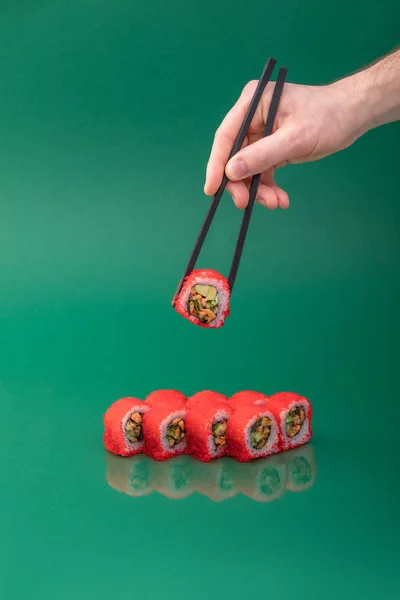 Рука держит красный суши-ролл с палочками для еды на зеленом фоне. набор рулонов — стоковое фото