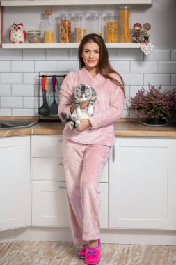 Evcil beyaz İskandinav mutfağında pijamalı genç bir kadın kediyi tutuyor.