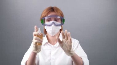 Tek kullanımlık eldivenli büyücü kadın ellerine antiseptik püskürtüyor. Tıbbi maskeli kadın, gri arka planda gözlüklü. 2019 Roman Coronavirus 2019-nCoV, COVID-19 konsepti.