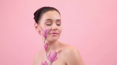 Yüzünde ve göğüs kafesinde boyalı manolya olan bir kadın. Vücudunda çiçek var. Pembe arkaplanda portre