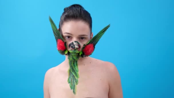 一个用鹦鹉翅膀化妆的女孩 身体艺术与鸟的脸 蓝色背景的女性模型 — 图库视频影像