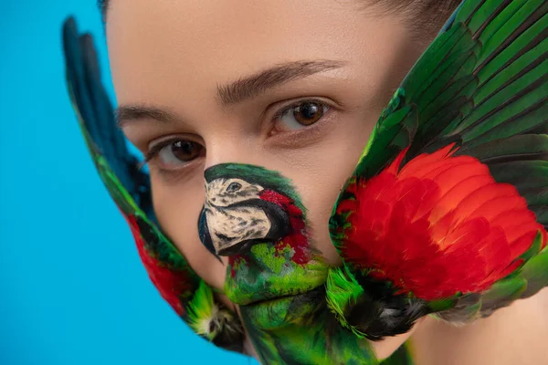 Woman with painted parrot bird on her face. Body art with real wings on the cheeks