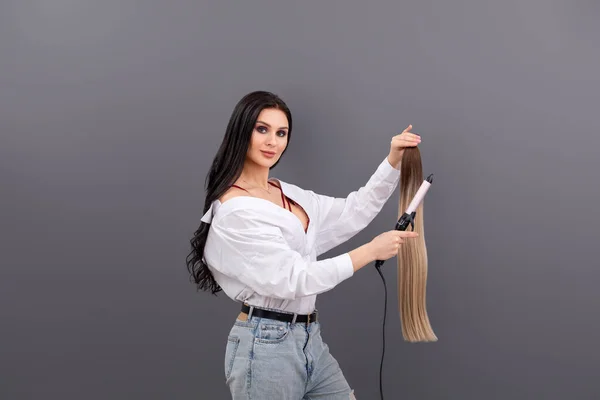 Fashion portrait of female hair artist or stylist holding sections of hair for extension with curling iron on grey background