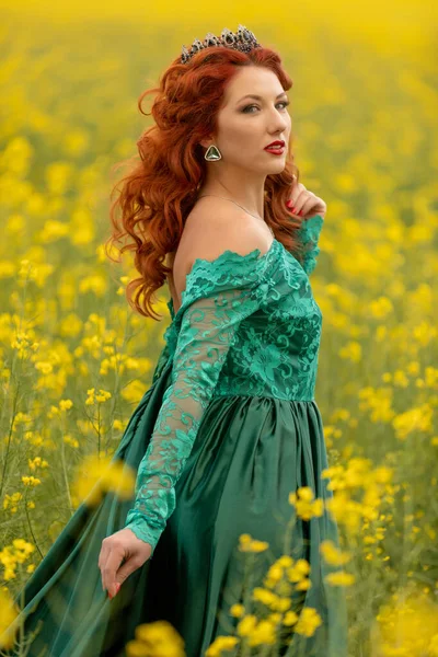 黄色の菜の花の分野で美しい赤毛の女性モデル 長い緑のドレスと王冠で身に着けている おとぎ話の姫や女王のイメージ ストック写真