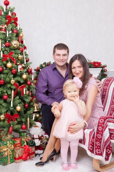 Celebración de Navidad o Año Nuevo. Feliz joven familia sentada en silla cerca del árbol de Navidad con regalos de Navidad. Una chimenea con medias de Navidad en el fondo — Foto de Stock