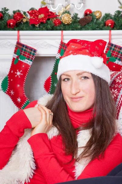 Celebração de Natal ou Ano Novo. Mulher nova em um jumper vermelho, colete de pele e chapéu de Santas, senta-se em uma cadeira em um interior de Natal, perto da lareira com meia de natal no fundo — Fotografia de Stock