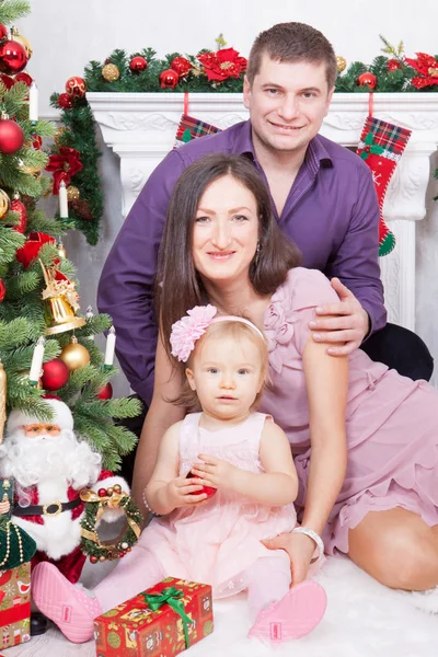 Celebración de Navidad o Año Nuevo. Feliz joven familia sentada en silla cerca del árbol de Navidad con regalos de Navidad. Una chimenea con medias de Navidad en el fondo — Foto de Stock