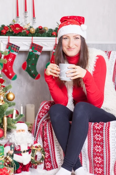 Celebração de Natal ou Ano Novo. Jovem mulher em um jumper vermelho, colete de pele e chapéu Santas, segura uma xícara na mão e sentado em uma cadeira em um interior de Natal, perto da lareira, árvore de Natal — Fotografia de Stock