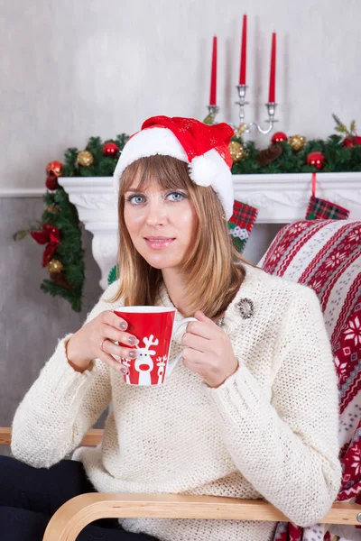 Celebração de Natal ou Ano Novo. Jovem mulher em um jumper de malha branca e chapéu Santas, segura uma xícara na mão e sentado em uma cadeira em um interior de Natal, perto da lareira, árvore de Natal — Fotografia de Stock