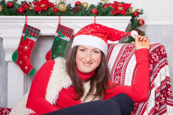 Celebração de Natal ou Ano Novo. Mulher nova em um jumper vermelho, colete de pele e chapéu de Santas, senta-se em uma cadeira em um interior de Natal, perto da lareira com meia de natal no fundo — Fotografia de Stock