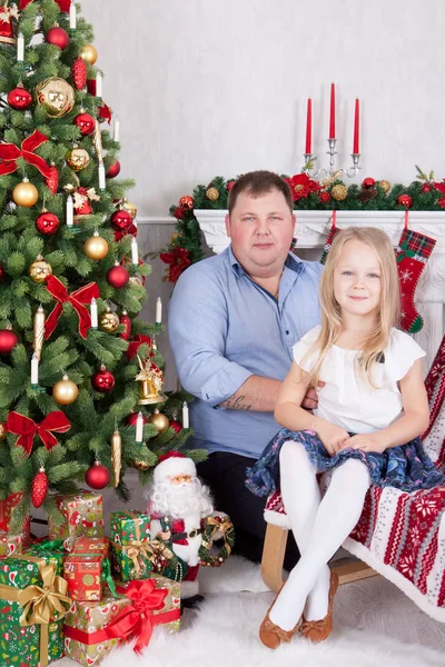Celebración de Navidad o Año Nuevo. Feliz padre e hija sentados en silla cerca del árbol de Navidad con regalos de Navidad. Una chimenea con medias de Navidad en el fondo — Foto de Stock