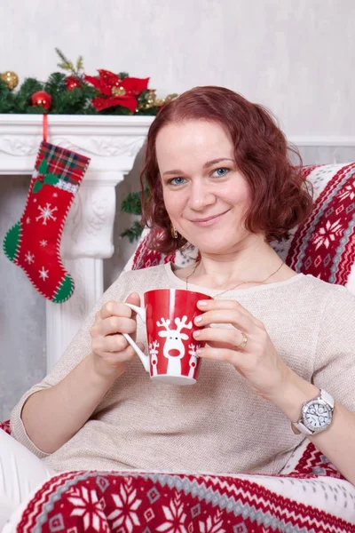 Celebração de Natal ou Ano Novo. Jovem mulher em um jumper de malha branca segura uma xícara na mão e sentado em uma cadeira em um interior de Natal, perto da lareira, árvore de Natal. Boas festas — Fotografia de Stock