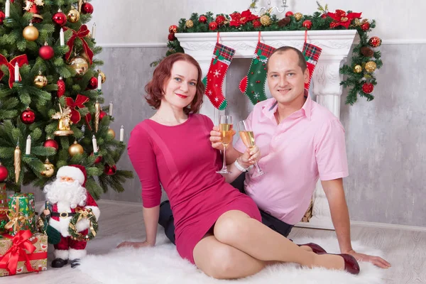 Celebração de Natal ou Ano Novo. Jovem casal bonito com uma taça de champanhe sentado perto da árvore de Natal com presentes de xmas. Uma lareira com meia de Natal no fundo. Boas festas — Fotografia de Stock