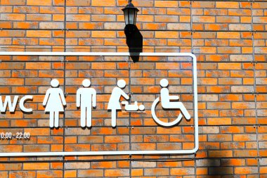 On the orange brick wall is a toilet signpost with white images of a man, a woman, a baby changing scene of a baby and a person in a wheelchair.