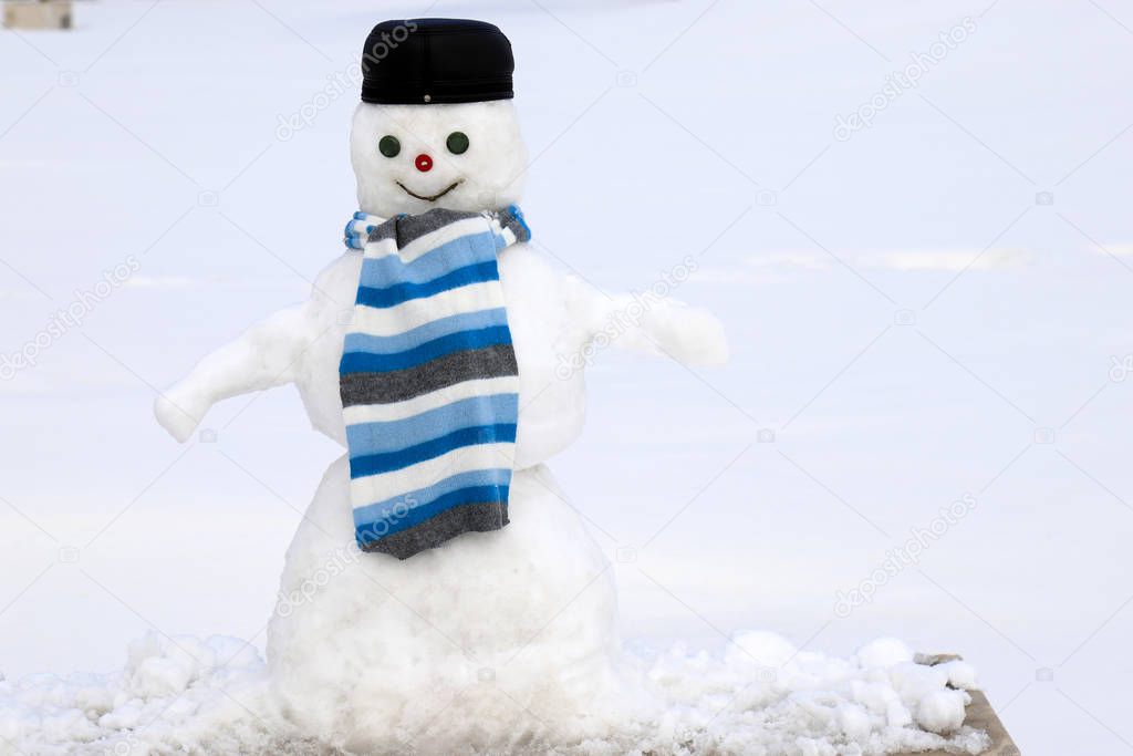 A Funny  white snowman in a black cap and a blue scarf is standing on a snowy meadow in winter, Christmas and New Year holidays, children entertainment and sports. 