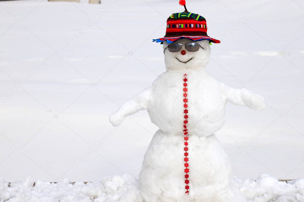 A Funny white snowman in a red cap is standing on a snowy meadow in winter, Christmas and New Year holidays, children entertainment and sports. 