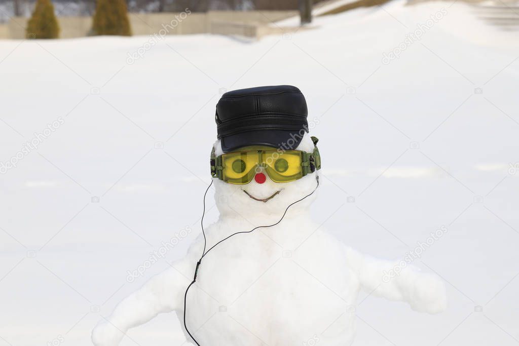 A funny smiling white snowman in a black cap, glasses and headphones stands in a snowy meadow in winter, on Christmas and New Year holidays. Children's winter entertainments and sports activities