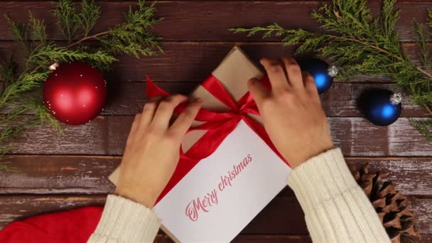 Top view hombre desenvolviendo regalos de Navidad con la inscripción Feliz Navidad en manos de escritorio de madera desde arriba — Vídeos de Stock