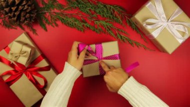 Üstten Görünüm Noel hediyelerini yukarıdan kırmızı zemin üzerine sarma eller. Hediye kahverengi kağıt sarılı ve mor kurdele bağlanmış