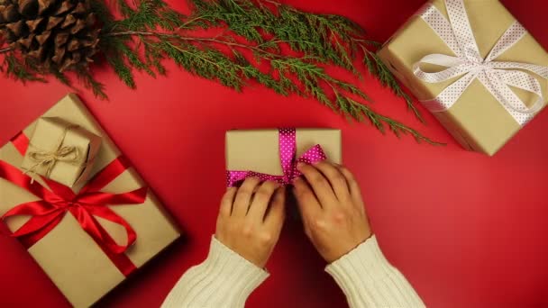 Руки зверху розпаковують різдвяні подарунки на червоному тлі зверху. Подарунок, загорнутий у коричневий папір і зав'язаний фіолетовою стрічкою — стокове відео