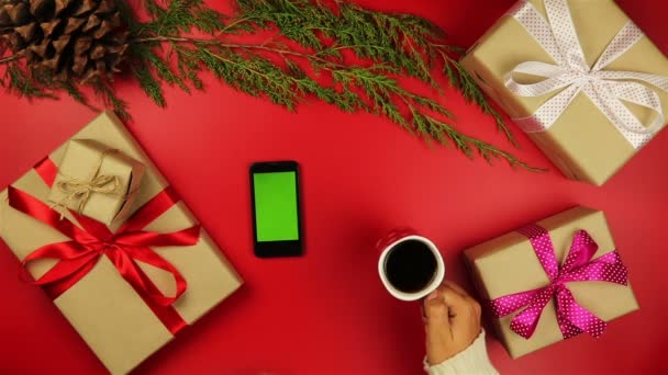 Top view christmas hands using touchscreen smartphone tablet christmas presents shopping list drinking coffee on red background from above. — Stock Video