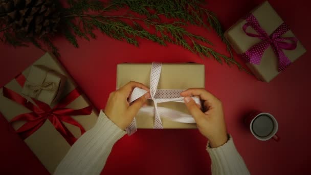 Το Top view χέρια περιτυλίγματος Χριστούγεννα παρουσιάζει σε κόκκινο φόντο από πάνω. Δώρο τυλιγμένο σε καφέ χαρτί και δεμένα λευκή κορδέλα — Αρχείο Βίντεο