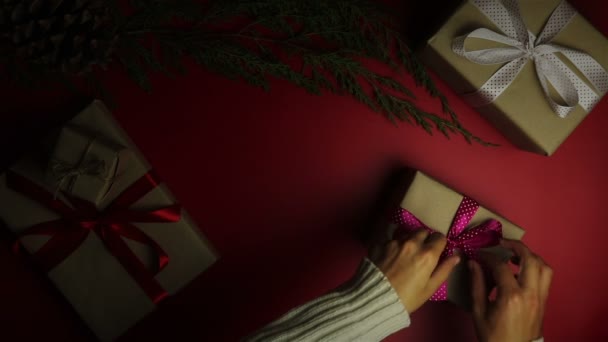 Las manos de vista superior que envuelven los regalos de Navidad en el fondo rojo desde arriba. Regalo envuelto en papel marrón y cinta púrpura atada — Vídeo de stock