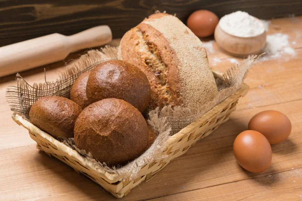 Assortment of fresh bread in a basket on a wooden table, composition with additional accessories — Stock Photo, Image