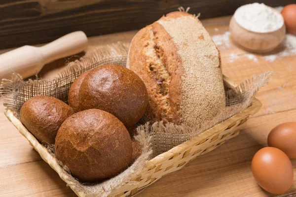 Assortment of fresh bread in a basket on a wooden table, composition with additional accessories — Stock Photo, Image