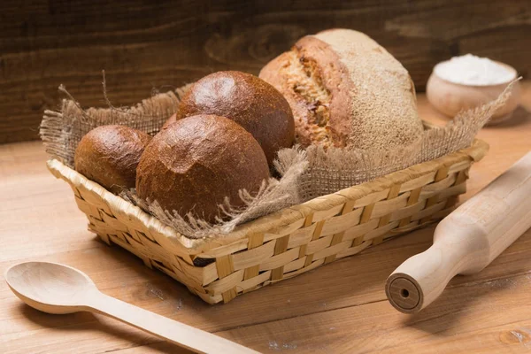Assortment of fresh bread in a basket on a wooden table, composition with additional accessories — Stock Photo, Image