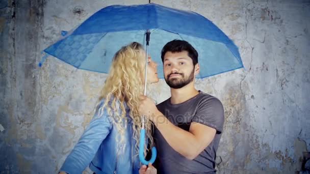 Ragazza baciare ragazzo sulla guancia sotto l'ombrello. Bella coppia giovane sotto l'ombrello blu. Ragazza bionda e uomo dai capelli scuri con un ombrello in cabina fotografica . — Video Stock