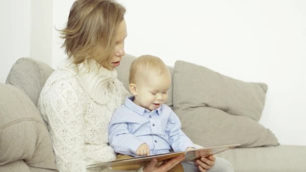 Omtänksam mor läser en bok och visar bilder till sin lille son, sitter i soffan hemma — Stockvideo