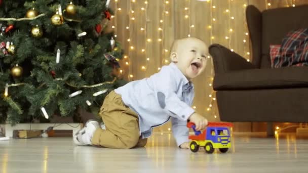 Kleiner aktiver blonder Junge, der mit einem Spielzeugauto auf dem Boden spielt, im Hintergrund ein Weihnachtsbaum und eine Girlande — Stockvideo