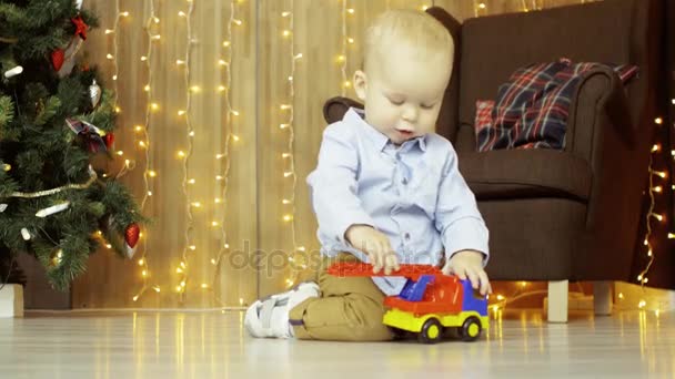 Χαριτωμένο Ευρωπαϊκό παιδί κυλά ένα αυτοκίνητο παιχνίδι στο πάτωμα, στο φόντο ένα χριστουγεννιάτικο δέντρο, πολυθρόνα και γιρλάντες — Αρχείο Βίντεο