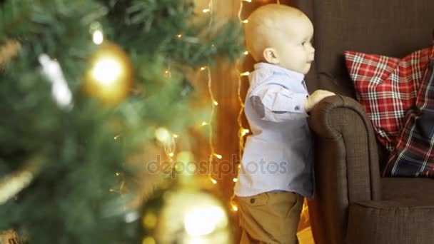 Bébé se tient sur le fauteuil avec ses mains, près de l'arbre de Noël, en arrière-plan une guirlande — Video